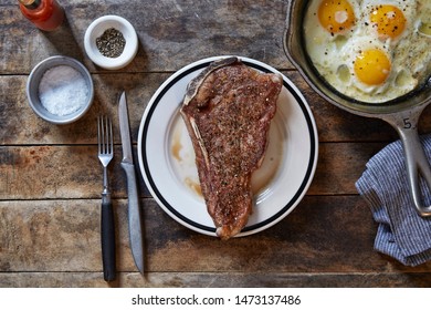 New York Strip Steak With Three Farm Fresh Eggs Served In  Cast-iron Skillet On Wooden Table With Tabasco Sauce And Salt And Pepper