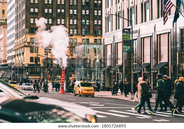 New York, USA - February\
8, 2020: Yellow taxis, steam stack and crowd of people on Fifth\
Avenue. Apple store on iconic shopping street. Pedestrians crossing\
urban road.