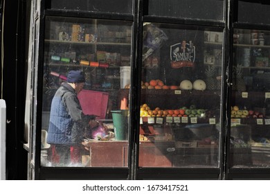 New York, USA / March, 2020 / An Isolated Grocery/shop Keeper Preparing Food For His Customers. An Array Of Fresh Food Can Be Seen Through The Shop Window, Framed By A Old Wooden Window Frame. 