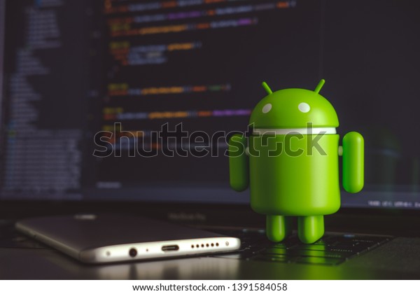 New\
York, USA - May 4, 2019 - Google Android figure standing on laptop\
keyboard next to mobile phone with code in\
background