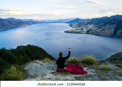 New Zealand, Wanaka Lake Travel Vacation. Happy Young Woman On The Mountain Top Hands Up With Wine Glass, Enjoying Mountain, Lake Landscape Outdoor View. Freedom, Happiness Lifestyle