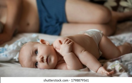 Newborn Baby Belly And Legs In Diaper, Lying On White Bed. Father Sleeping On Bed With Little Baby Boy Or Girl.