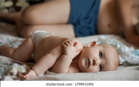 Newborn Baby Belly And Legs In Diaper, Lying On White Bed. Father Sleeping On Bed With Little Baby Boy Or Girl.