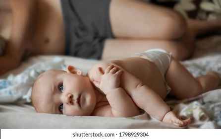 Newborn Baby Belly And Legs In Diaper, Lying On White Bed. Father Sleeping On Bed With Little Baby Boy Or Girl.