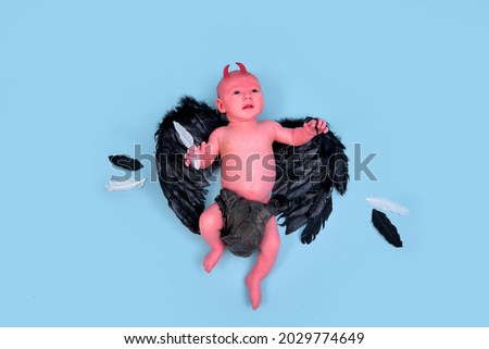 A newborn baby boy with black demon wings on a blue studio background, copy space. An infant caucasian child in a diaper aged one month