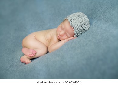 Newborn Baby Boy Sleeping In The Fetal Position On A Blue Background In A Knitted Cap.