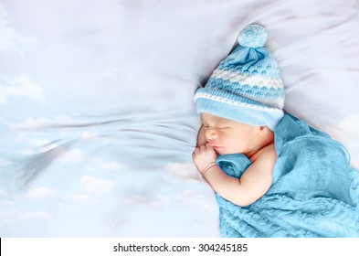 Newborn Baby Boy Sleeping On White Silk Bed Floating Clouds Wearing Blue Hat And Blanket Isolated In White Background