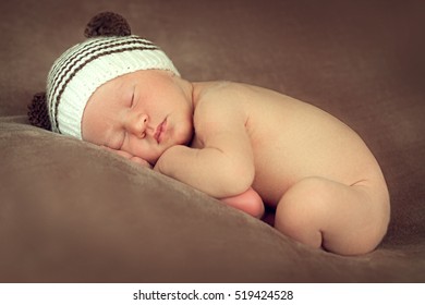 Newborn Baby Boy Sleeping Peacefully In The Fetal Position On A Brown Background With The Beanie With Ears In The Image Of Teddies