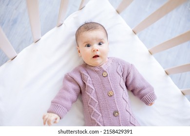 A Newborn Baby In A Crib In A Brown Warm Jumpsuit On A White Cotton Bed Has Woken Up In The Morning Or Is Falling Asleep, A Surprised Portrait Of A Baby