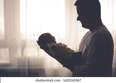 Newborn Baby And Dad, A Newborn Baby In Her Husband's Arms, A Baby In A Man's Arms. Dad And Son, Fatherhood Concept. Real Room, Dark Style And Toning