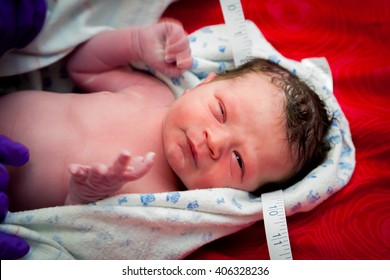 A Newborn Baby Gets His Head Measured By The Midwife.  He Has One Eye Opened And The Other Partially Open.  His Hands Are Moving Quickly, But His Head Is Still.