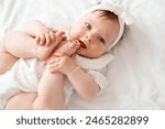 newborn baby girl plays with her legs pushing them into her mouth in white clothes on the bed at home on her back, top view, baby with legs in her mouth, space for text