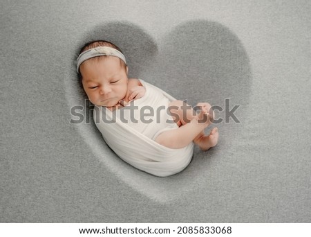 Newborn baby girl sleeping in heart shape form swaddled in white fabric. Adorable infant child kid napping during studio photoshoot