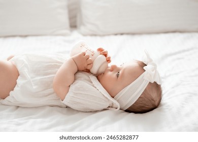 Newborn baby girl sucks a bottle of milk in white clothes on the bed at home on her back, cute baby eats, space for text.