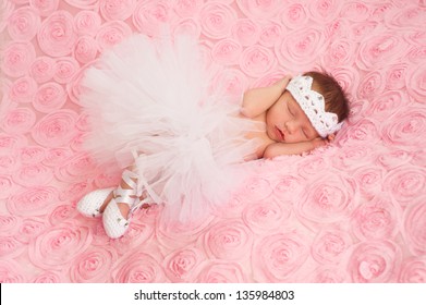 Newborn Baby Girl Wearing A White Crocheted Crown, Ballerina Tutu, And Ballet Slippers. She Is Sleeping On Pink Rose Ribbon Fabric.