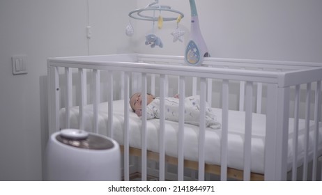 A newborn baby lies in a crib against the background of an ultrasonic humidifier.