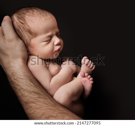 Newborn Baby lying on Father Hand over Black Background. Child sleeping in Fetal Embryo position. Small Kid artistic Portrait. Baby Care and Parenting Concept