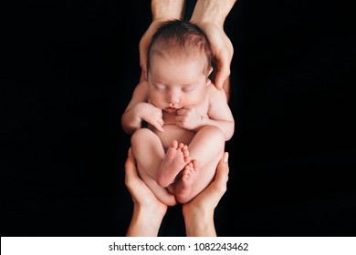 Newborn Baby Lying On The Hands Of Parents On A Black Background. Imitation Of A Baby In The Womb. Beautiful Little Girl Sleeping Lying On Her Back