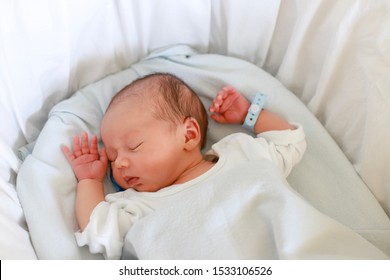 Newborn Baby With Name Tag Bracelets Sleeping In Bassinet Bed At Hospital. Mixed Race Asian-German Infant First Day Of Life.