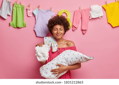 Newborn Baby Rests In Mothers Hands. Pleased Curly Haired Woman Caring Mom Holds Sleeping Baby Wrapped In Blanket On Hands Has Diaper Bodysuit On Shoulder Poses Indoor. Happy Family Concept.