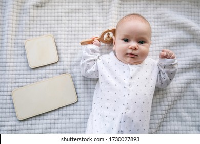 A newborn baby of two months old with a wooden natural rattle in his hand on a white bedspread. Empty labels for text