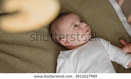 Newborn boy gazes at hanging mobile while reaching hand on knitted blanket in nursery; gentle curiosity.