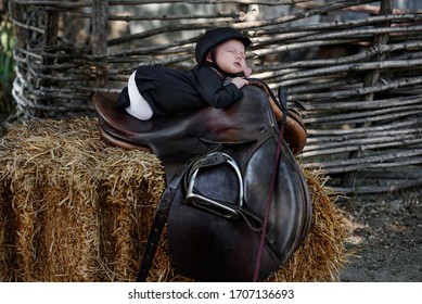 A Newborn Child In Equestrian Clothes Lies Sleeping On A Saddle In The Hay Against The Background Of A Fence
