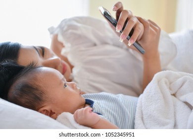 Newborn Concept. Mother And Child On A White Bed. Mom And Baby Boy Playing In Bedroom. Mother Is Playing Mobile Phone On Bed.