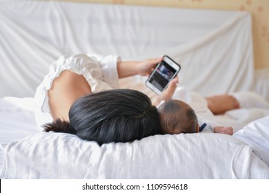 Newborn Concept. Mother And Child On A White Bed. Mom And Baby Boy Playing In Bedroom. Mother Is Playing Mobile Phone On Bed.