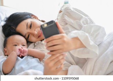 Newborn Concept. Mother And Child On A White Bed. Mom And Baby Boy Playing In Bedroom. Mother Is Playing Mobile Phone On Bed.