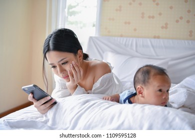 Newborn Concept. Mother And Child On A White Bed. Mom And Baby Boy Playing In Bedroom. Mother Is Playing Mobile Phone On Bed.