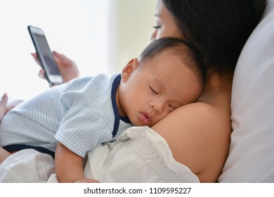 Newborn Concept. Mother And Child On A White Bed. Mom And Baby Boy Playing In Bedroom. Mother Is Playing Mobile Phone On Bed.