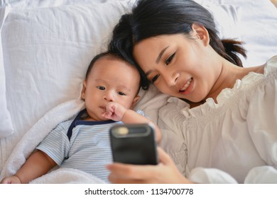 Newborn Concept. Mother And Child On A White Bed. Mom And Baby Boy Playing In Bedroom. Mother Is Playing Mobile Phone On Bed.