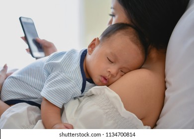 Newborn Concept. Mother And Child On A White Bed. Mom And Baby Boy Playing In Bedroom. Mother Is Playing Mobile Phone On Bed.