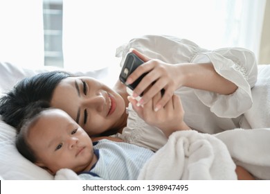 Newborn Concept. Mother And Child On A White Bed. Mom And Baby Boy Playing In Bedroom. Mother Is Playing Mobile Phone On Bed.