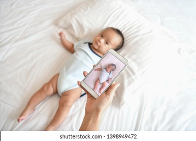 Newborn Concept. Mother And Child On A White Bed. Mom And Baby Boy Playing In Bedroom. Mother Is Playing Mobile Phone On Bed.