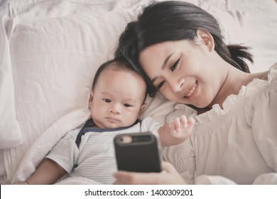 Newborn Concept. Mother And Child On A White Bed. Mom And Baby Boy Playing In Bedroom. Mother Is Playing Mobile Phone On Bed.