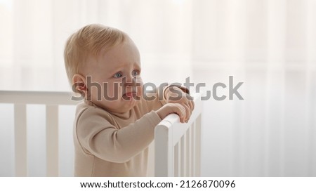 Newborn crying baby portrait. Little infant tired and hungry, start crying standing in crib, looking aside at empty space in nursery, panorama