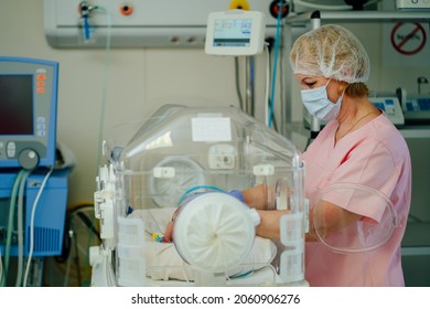 Newborn Is Placed In The Incubator. Neonatal Intensive Care Unit. Nurse Standing Near Hospital Bed With A Baby Preparing It For Treatment.