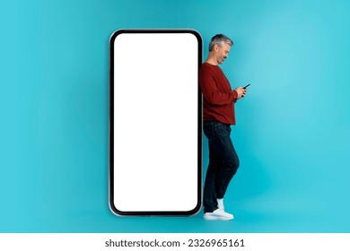 Newest mobile app concept. Cheerful happy handsome grey-haired middle aged man 40s in casual outfit using cell phone, standing by big phone with white blank screen, blue background, mockup - Powered by Shutterstock