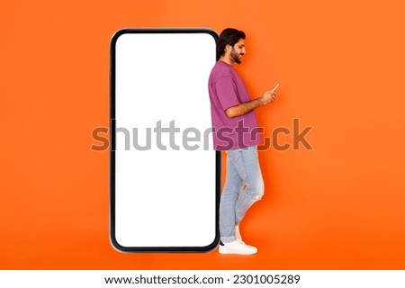 Newest mobile app. Cool stylish handsome young eastern guy standing by big smartphone with white empty screen, using cell phone and smiling, mockup, orange studio background, copy space