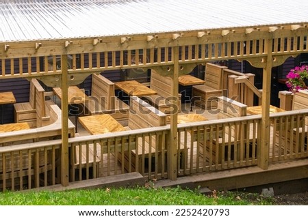 A newly built rustic outdoor dining area, known as a veranda or deck with wooden benches, tables, and wooden walls. The rustic restaurant balcony has an empty seating area with a pergola overhead.