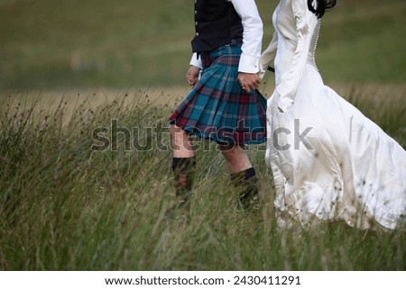 A newly married Scottish couple strolls through a grassy field in Glencoe, the Scottish highlands. He dressed in a Scottish Kilt and she dressed in a white wedding dress