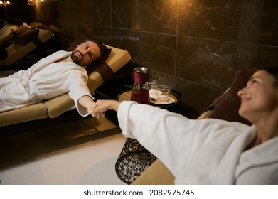 Newlywed, beautiful couple of a handsome attractive man in white bathrobe lying on sun lounger and looking at his beloved partner holding her hand in his hand, enjoying honeymoon at health spa resort - Powered by Shutterstock