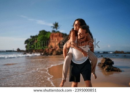 Newlyweds celebrate love with piggyback fun on sandy beach. Honeymoon joy, playful couple near sea. Romance, vacation in tropics. Bride, groom enjoy sunset. Exotic island getaway, love, happiness.