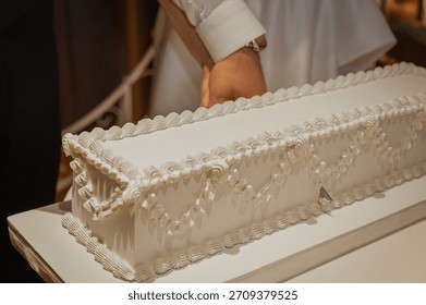 Newlyweds cutting a long rectangular white wedding cake together during reception. Bride and groom hands holding a knife. Rustic venue with pottery background