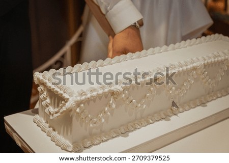 Newlyweds cutting a long rectangular white wedding cake together during reception. Bride and groom hands holding a knife. Rustic venue with pottery background