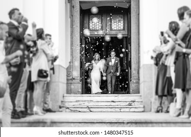 Newlyweds exiting the church after the wedding ceremony, family and friends celebrating their love with the shower of soap bubbles, custom undermining traditional rice bath. Black and white.