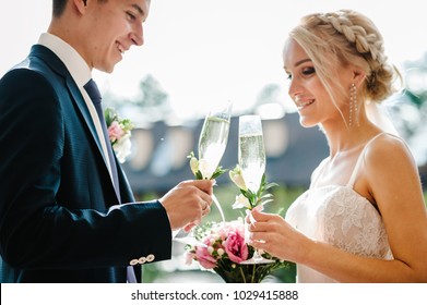 Newlyweds Holding Glasses Of Champagne For Toast In Hands Of The Outdoor. Standing On Wedding Ceremony Under The Arch Decorated With Flowers And Greenery In The Backyard Banquet Area.