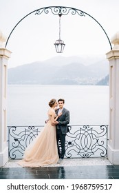 Newlyweds hug and almost kiss under an old arch against the backdrop of Lake Como